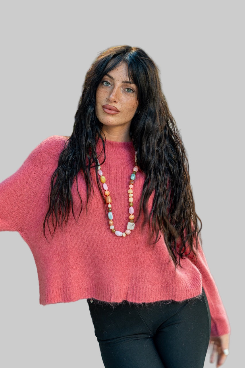 Woman wearing a pink sweater and black pants with a long necklace, standing outdoors.